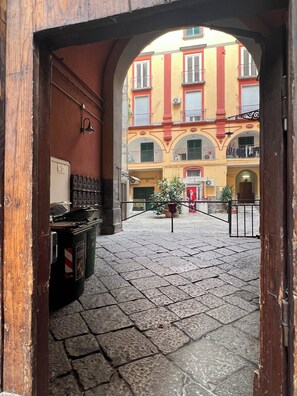 An arched doorway leading to a courtyard with a tiled floor, a row of trash bins, and a building with balconies in the background. - New Listing (Napoli)