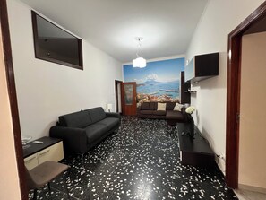 A hallway with a black and white speckled floor, a black sofa, a small table with a lamp, and a wall-mounted TV. - New Listing (Napoli)