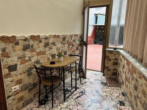 Una habitación con paredes de piedra, una mesa y sillas de madera, una planta en maceta y vistas a un espacio exterior con suelo rojo y un edificio al fondo.