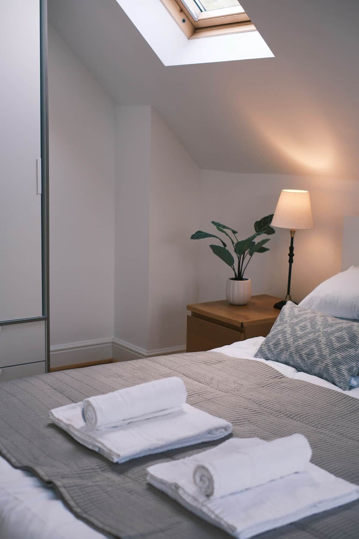 Un dormitorio moderno con tragaluz, lámpara de noche, planta en maceta y toallas ordenadas sobre la cama.