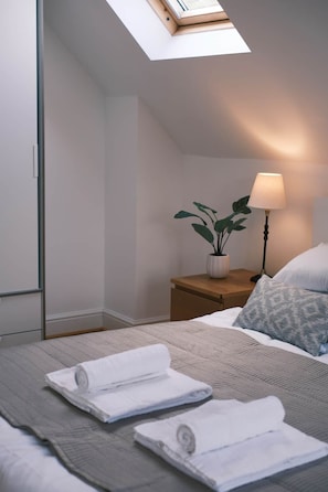 Une chambre à coucher moderne avec un puits de lumière, une lampe de chevet, une plante en pot et des serviettes soigneusement pliées sur le lit.