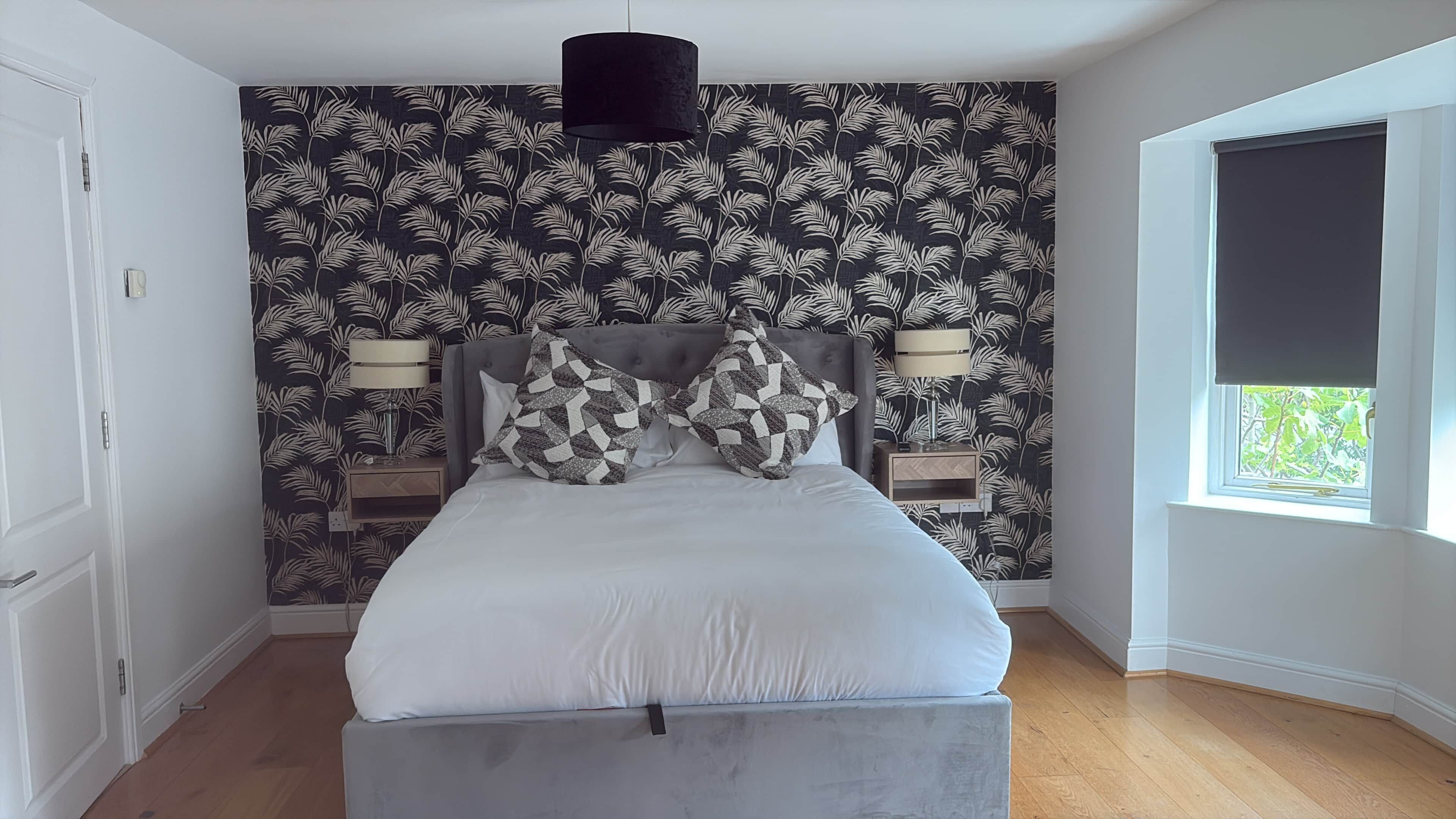 Ein Schlafzimmer mit einem Bett, zwei Nachttischlampen, einem Wandpapier mit Muster und einem Fenster mit Blick.