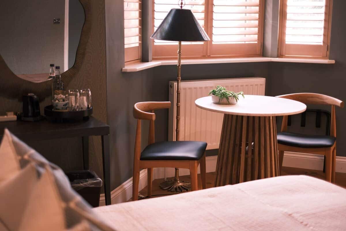 A bedroom with a bed, a round table with chairs, a lamp, and a window with blinds.