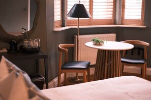 Une chambre à coucher avec un lit, une table ronde entourée de chaises, une lampe et une fenêtre avec des stores.