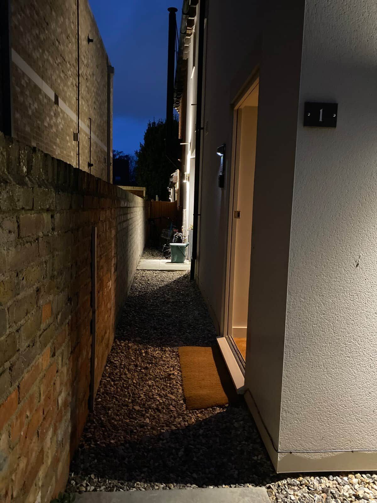 A narrow alleyway with a brick wall on one side and a white building on the other. There is a door with a number plate, a small mat at the entrance, and a gravel path.