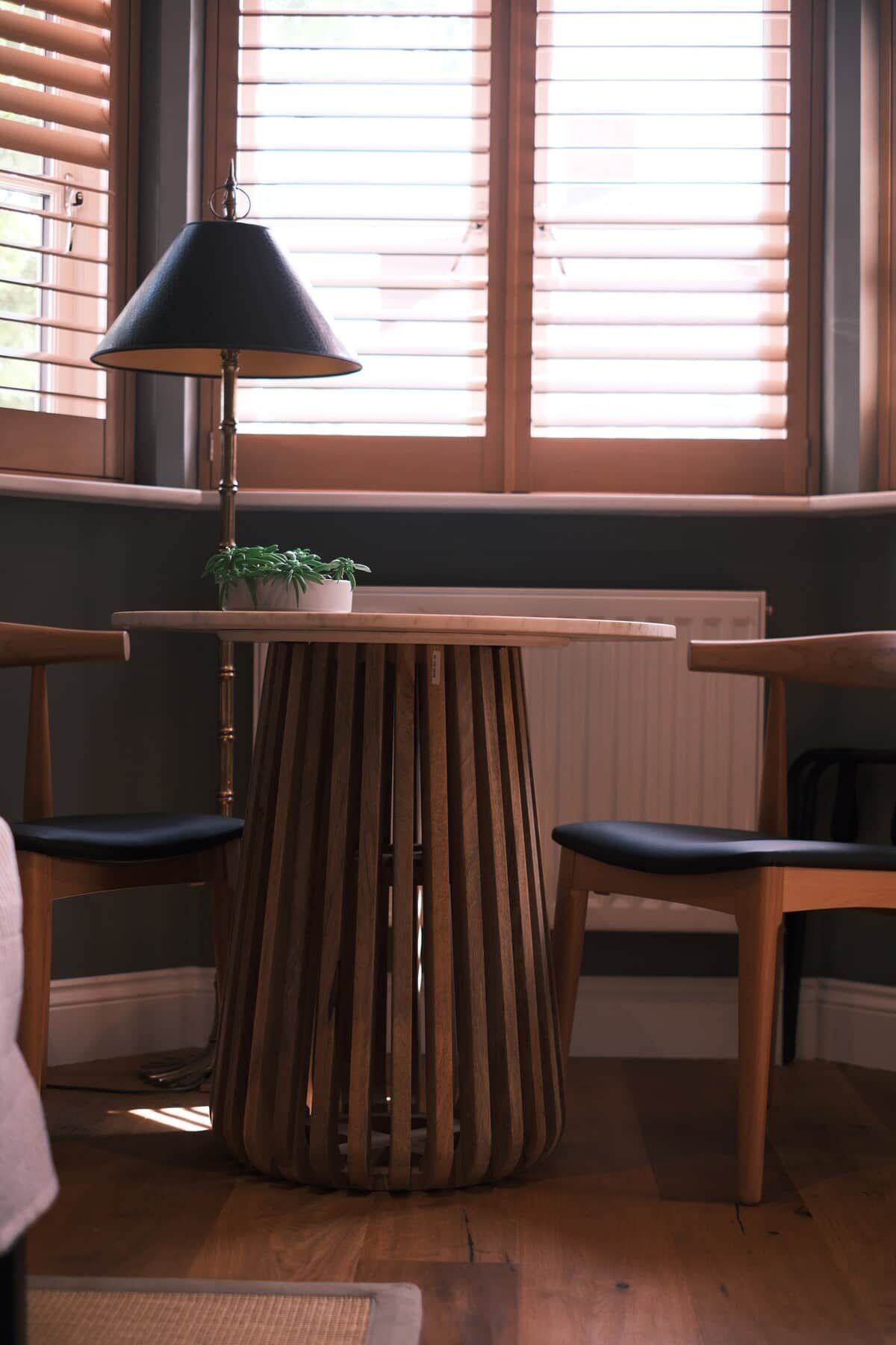 A room with a wooden table and two chairs, a lamp, and a window with blinds.