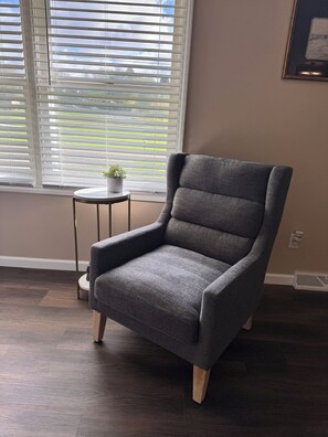 Een grijze fauteuil met houten poten, geplaatst voor een raam met jaloezieën, een klein rond tafeltje met een potplant en een lijst met een afbeelding aan de muur.