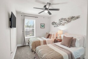 Quarto com duas camas, ventilador de teto, TV, janela com persianas e decoração na parede.