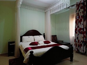 Un lit avec une tête de lit en bois foncé, une literie blanche et des coussins rouges. Il y a une petite table de chevet avec une bouteille et une télécommande. Les murs de la pièce sont d’un vert clair et la fenêtre est ornée d’un rideau à motifs.