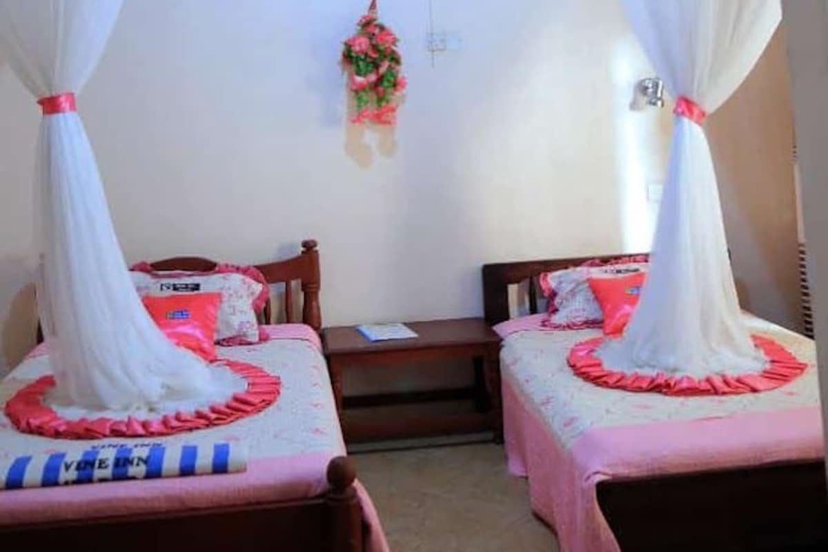 A room with two beds, each with a canopy. There is a small table and a chair, and a decorative hanging in the center.