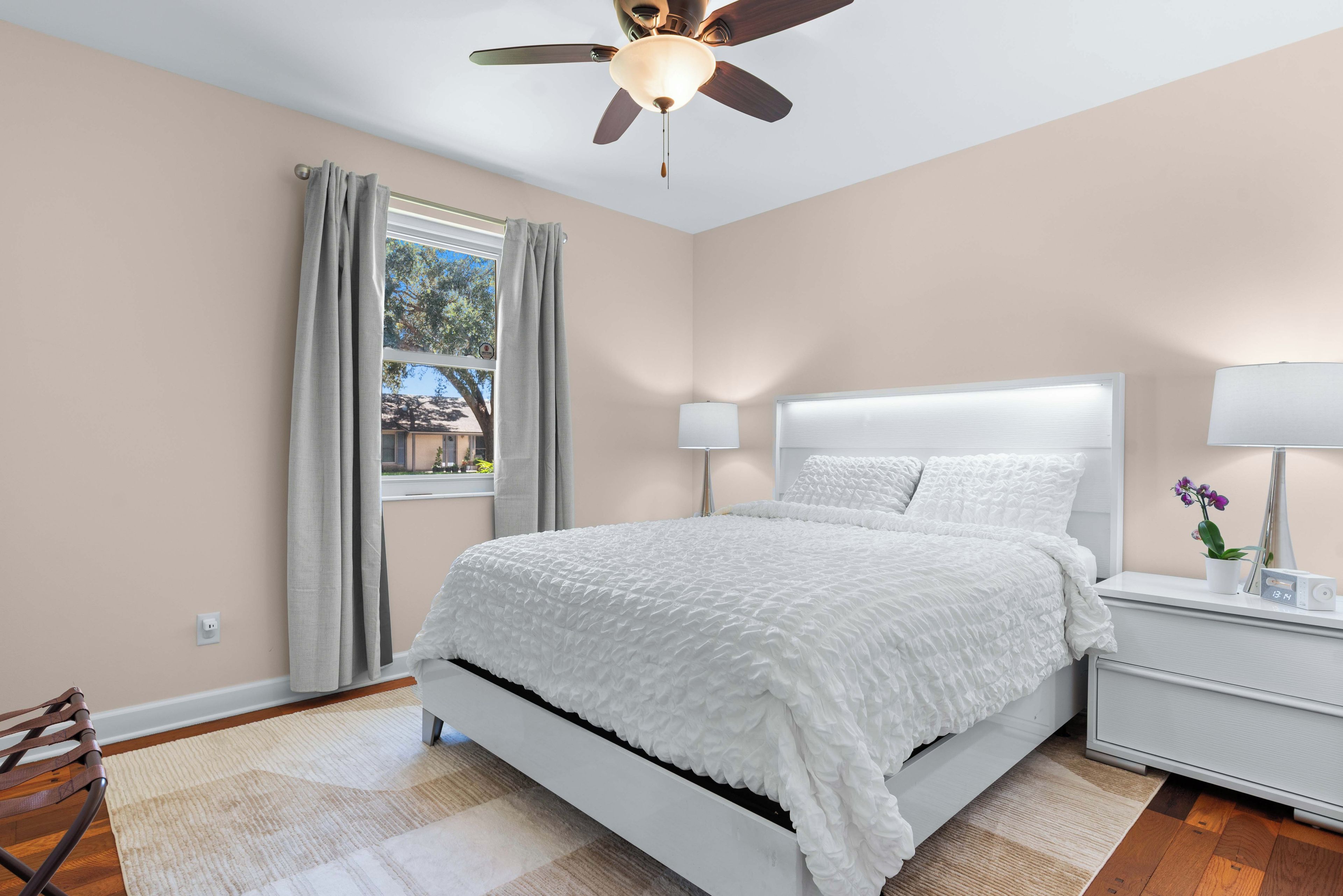 A bedroom with a bed, a ceiling fan, a window with curtains, a bedside lamp, and a nightstand with a clock and a plant.