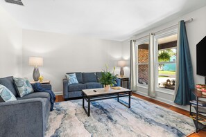 A living room with a sofa, a coffee table, and a TV. There is a window with curtains and a view of the outside.
