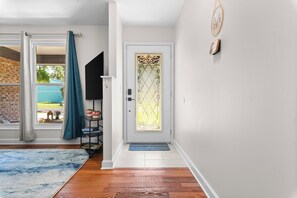 A hallway with a door, a window with curtains, a small table with plants, and a rug.