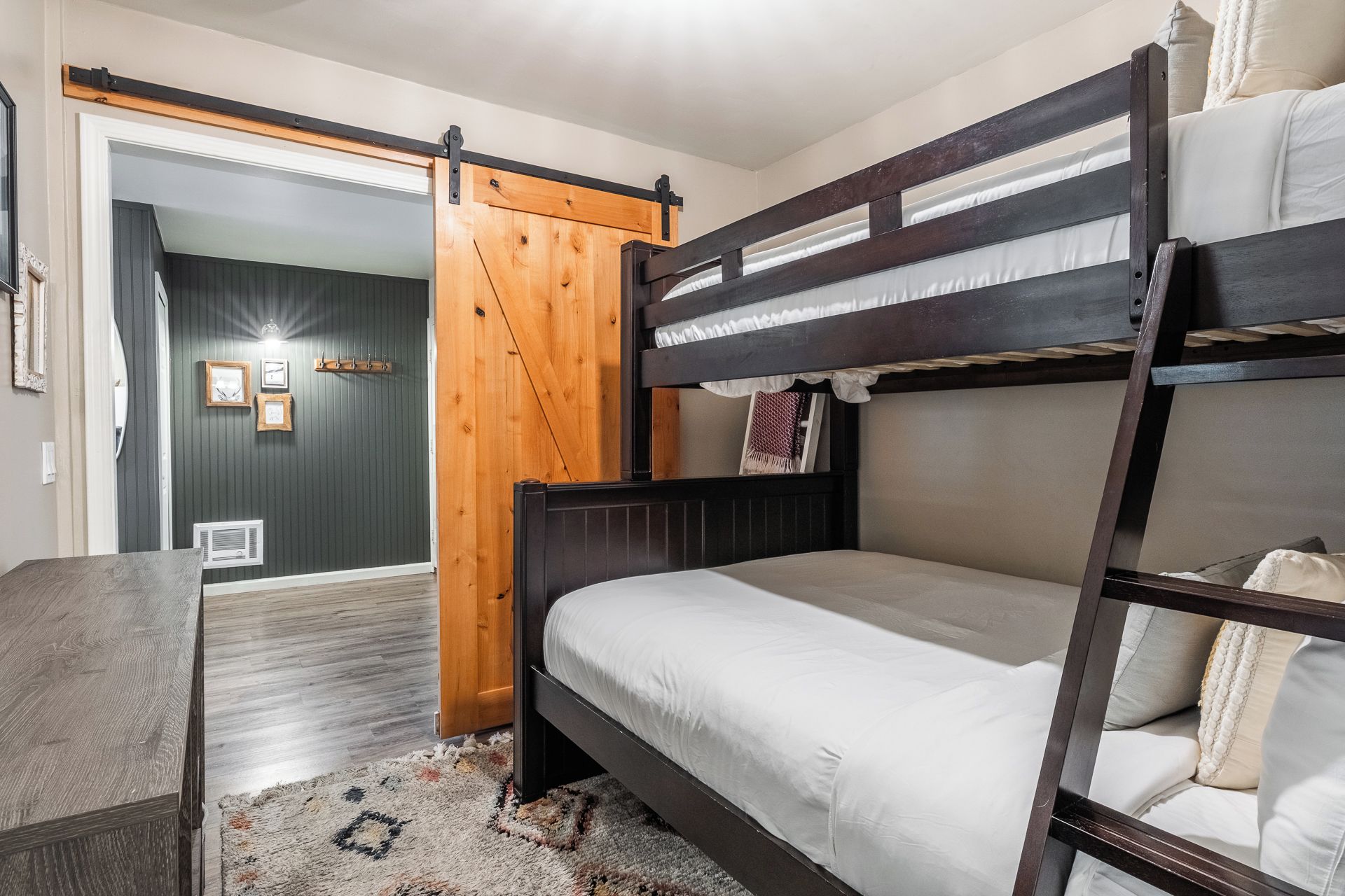 A bunk bed with a wooden frame and white bedding, a wooden door with a sliding mechanism, and a small table with a patterned rug.