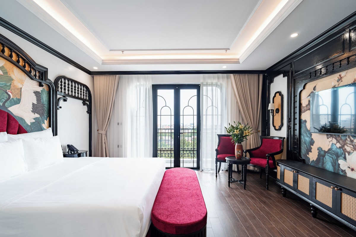 Quarto de hotel com uma cama grande, um banco vermelho, uma escrivaninha com cadeira e uma televisão.