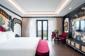 Quarto de hotel com uma cama grande, um banco vermelho, uma escrivaninha com cadeira e uma televisão.
