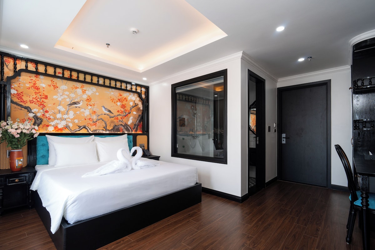 Quarto de hotel moderno com uma cama grande, uma obra de arte na parede, um espelho e uma porta que dá para outro cômodo.