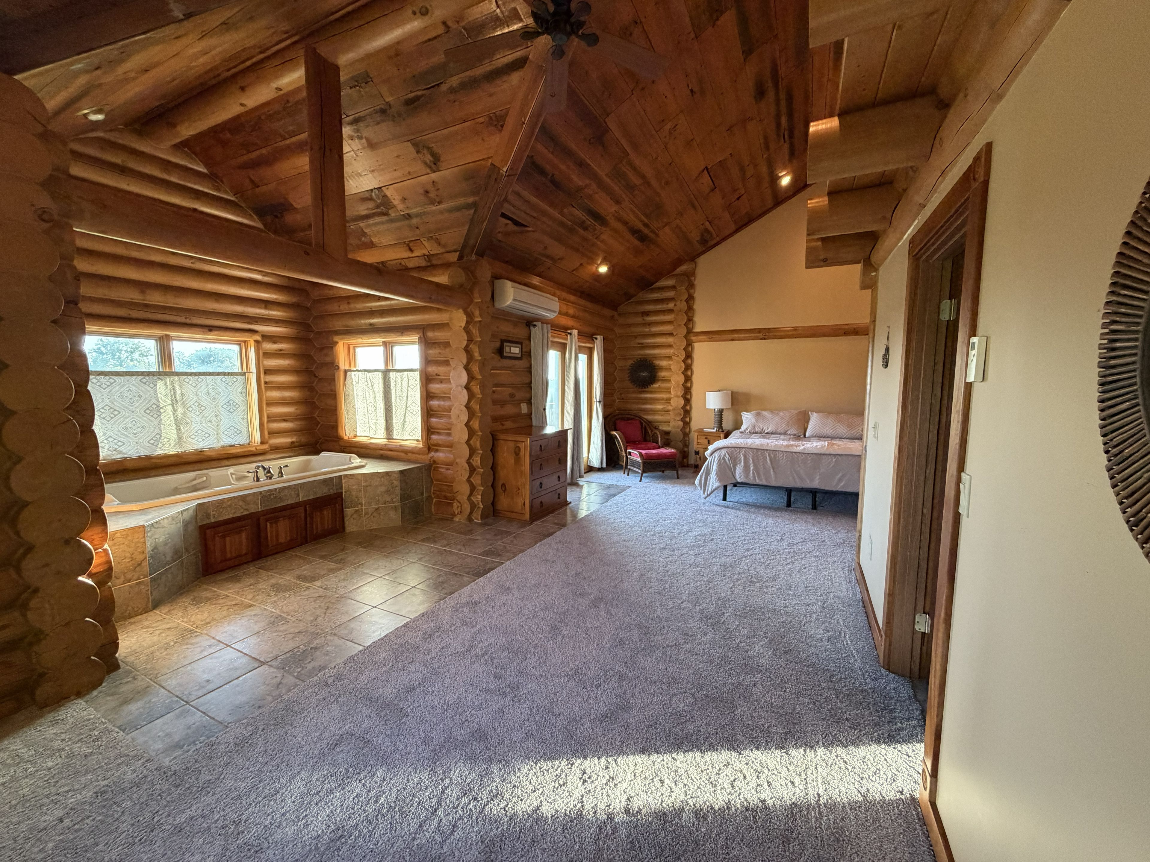 A log cabin bedroom with a large bed, a sitting area with a chair, a window, and a built-in bathtub.