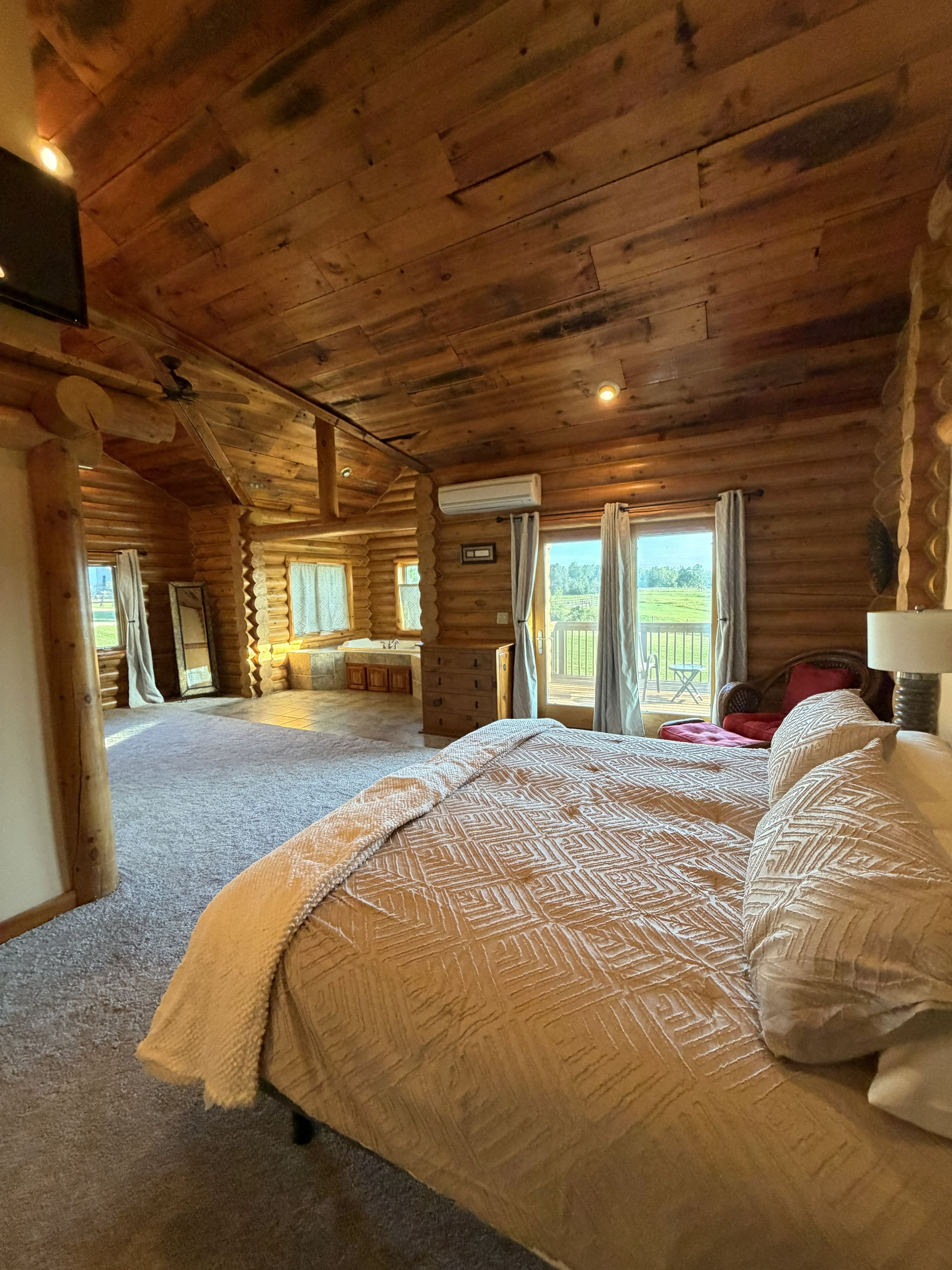Quarto de cabana de madeira com uma cama grande, teto de madeira e vista para uma paisagem verde pela janela.