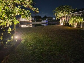 A nighttime view of a residential area with a grassy lawn, a small pond, and a house with a covered porch. - Ultimate Waterfront Escape: Pool, Kayaks & Fun! (Cape Coral)