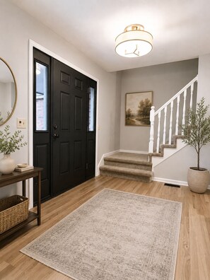 A hallway with a dark door, a staircase, a light fixture, a framed picture, a potted plant, and a woven basket. - Luxury Furnished Home - 90 Day Minimum Stay (Lincoln)