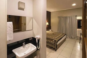 Ein Hotelzimmer mit Bad, das ein Waschbecken, einen Spiegel und einen Föhn bietet. Das Zimmer verfügt über ein Bett mit einem gemusterten Überwurf und einen Vorhang.
