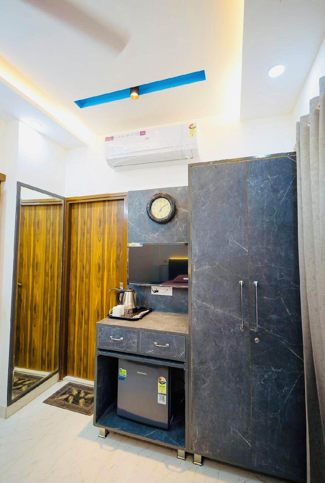 A compact room with a wardrobe, a small kitchenette, a clock, and a ceiling light fixture.