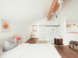 A bedroom with a bed, a chair, a bedside table, a wooden bench, a suitcase, and a shower area. - Luna Mia 1/chalet (Schwangau)