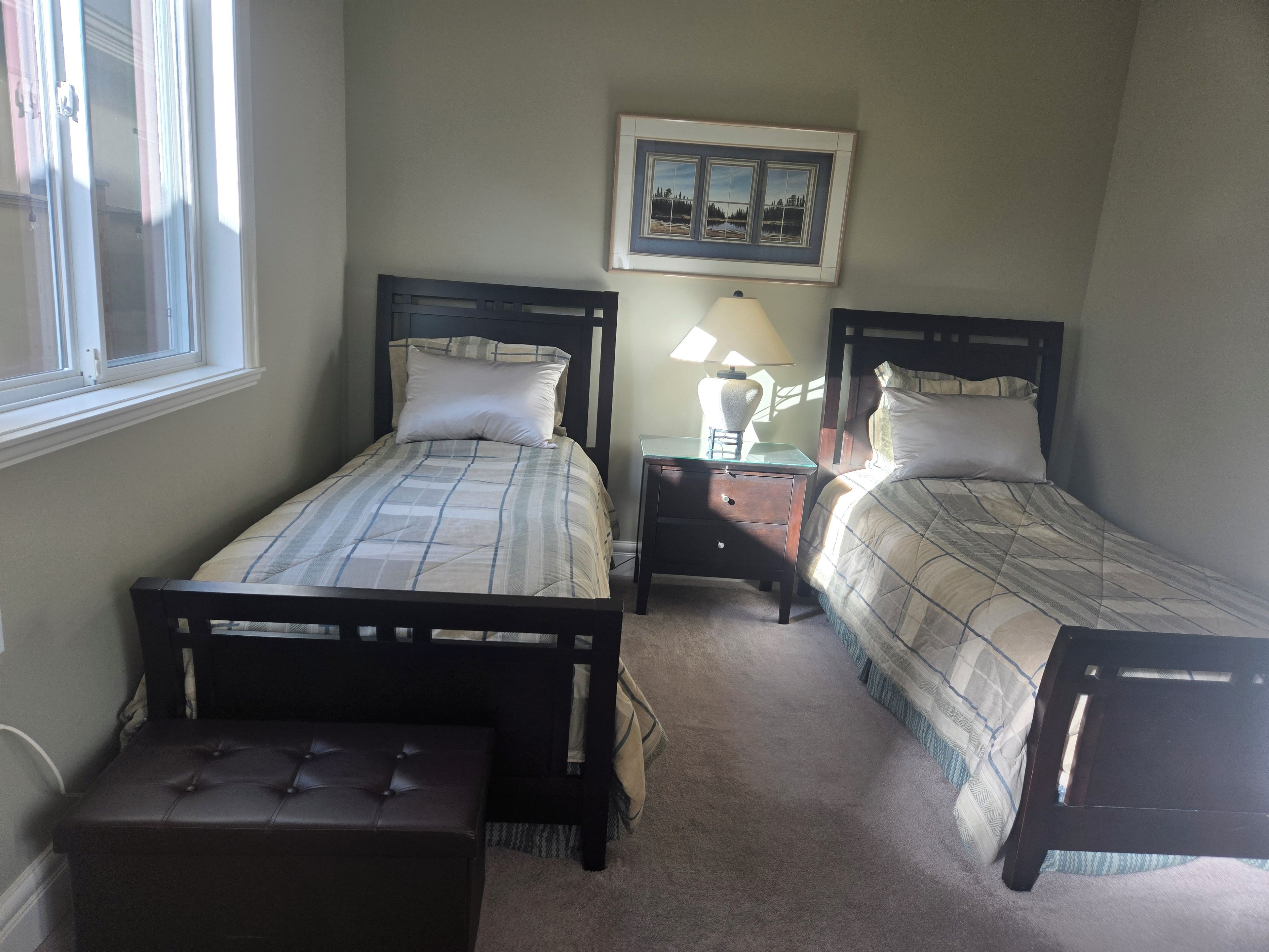 A bedroom with two single beds, a nightstand with a lamp, a bench, and a framed picture on the wall.