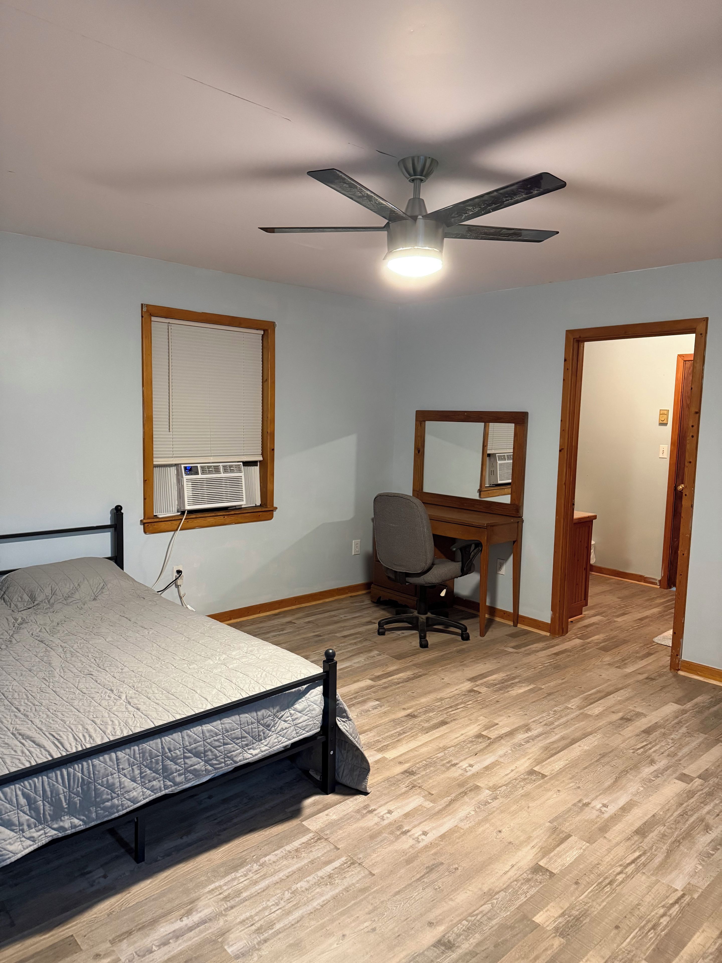 Un dormitorio con cama, escritorio, silla, ventilador de techo y ventana con persianas.