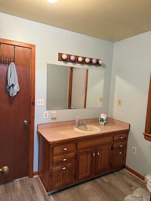 Un bagno con un mobiletto vanità in legno, un lavandino, uno specchio illuminato, un porta asciugamani e una porta.