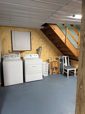 A laundry room with two washing machines, a drying rack, a chair, and a window with blinds.