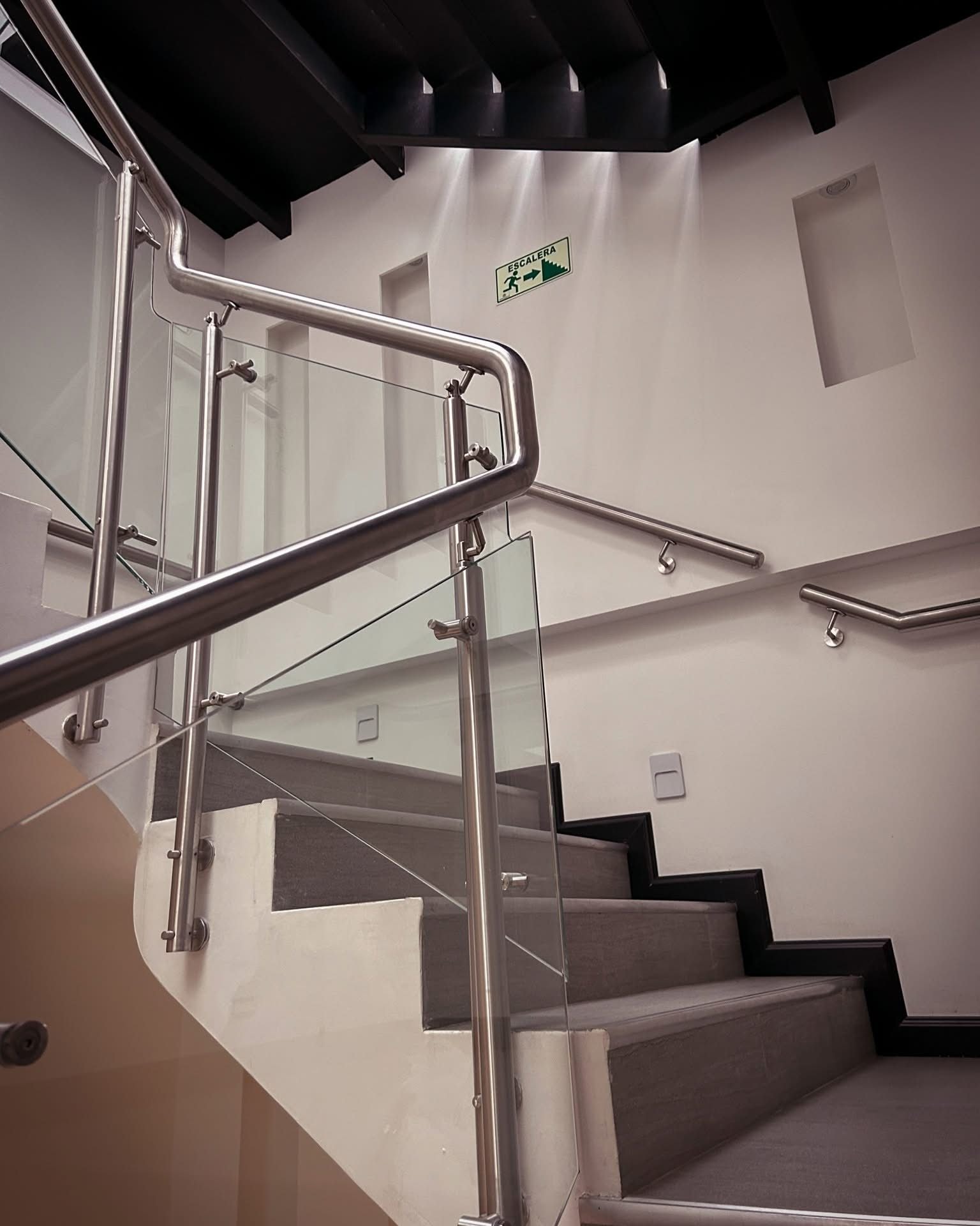 A staircase with metal handrails and glass panels, leading to a landing with a sign indicating an emergency exit.