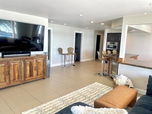 A modern living room with a flat-screen TV, wooden cabinet, seating area, and a kitchen in the background. - Lakefront, Private Dock & Hot Tub - Sleeps 14 (Moses Lake)
