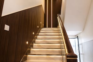 Una escalera con un lateral de pared de madera y otro de barandilla de cristal, con escalones iluminados.
