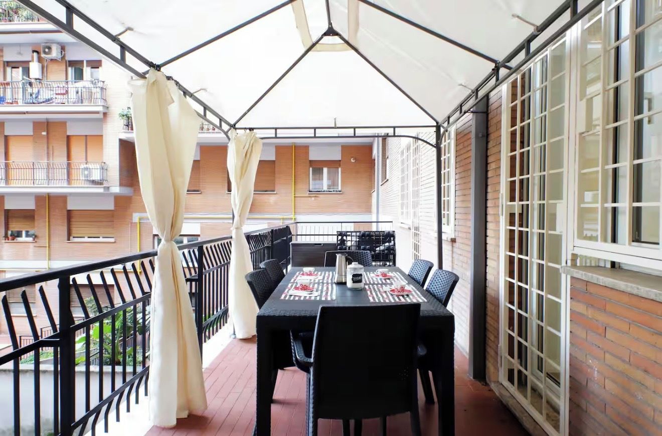 Un balcone con tavolo e sedie, un tendaggio bianco e vista su un edificio con finestre.