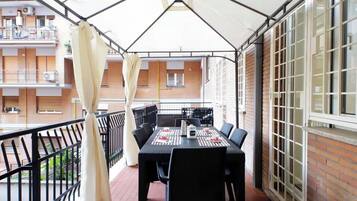 Un balcone con tavolo e sedie, un tendaggio bianco e vista su un edificio con finestre.