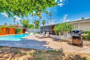 Un giardino sul retro con piscina, lettini da relax, barbecue e una casa con recinzione bianca.