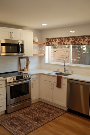 A modern kitchen with white cabinetry, stainless steel appliances, and a window with a view of a wooden fence. - Private 2 Bed ADU Near Airport & Hospitals (Kent)