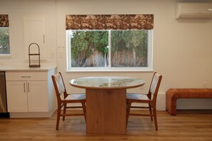 Een eetgedeelte met een ronde tafel, twee stoelen, een bank, een raam met uitzicht op bomen, en een keuken met kasten en een spoelbak.