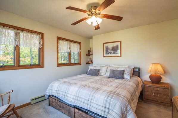 Un dormitorio con cama, ventilador de techo, mesita de noche, lámpara, silla y dos ventanas con cortinas.