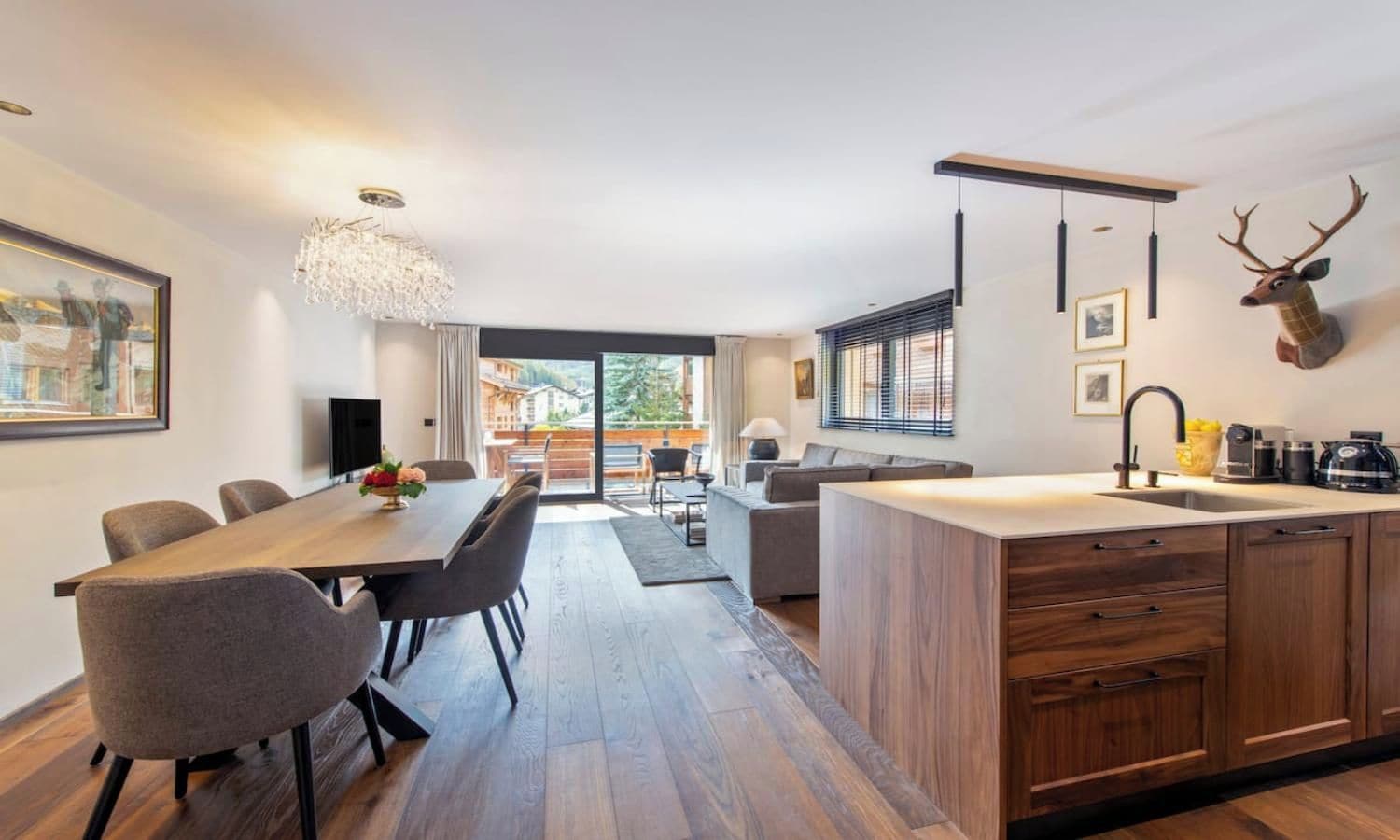 A modern kitchen with a central island, dining area with chairs, and a large window with blinds.