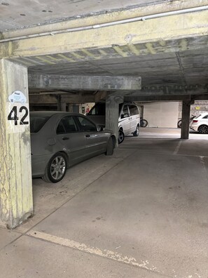 Autotalli, jossa on betonipilarit, ja sen alla on parkkeerattu harmaa ja valkoinen auto.