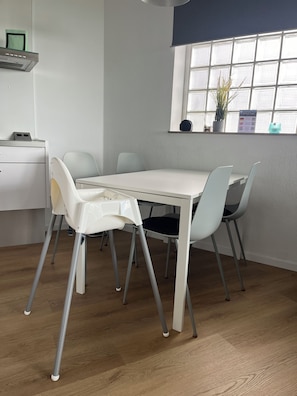 Un comedor con una mesa y sillas blancas, una ventana con persianas, un estante con una planta y un pequeño escritorio con una lámpara.