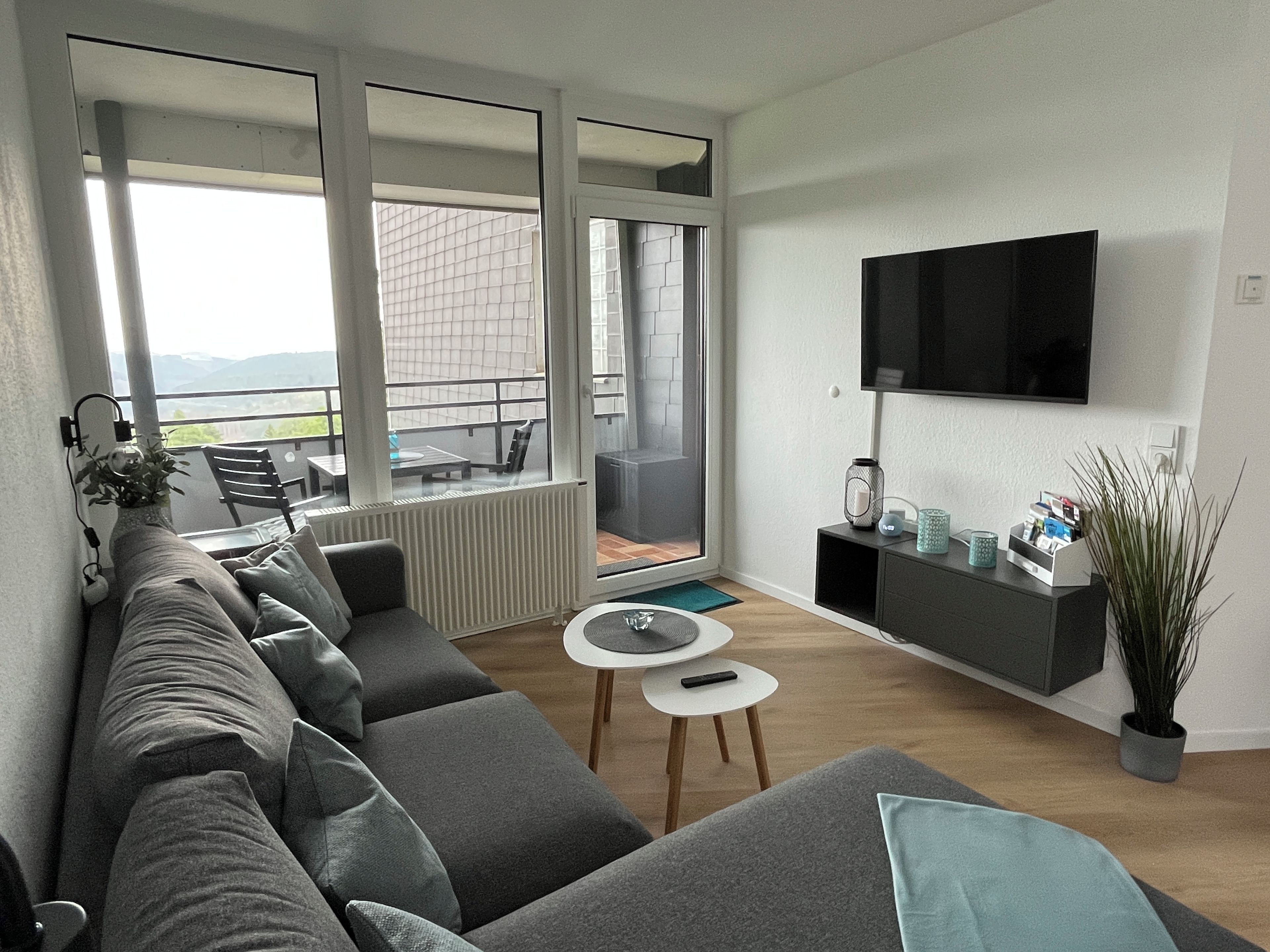 A modern living room with a grey sofa, a small round table, a mounted TV, and a balcony with a view.