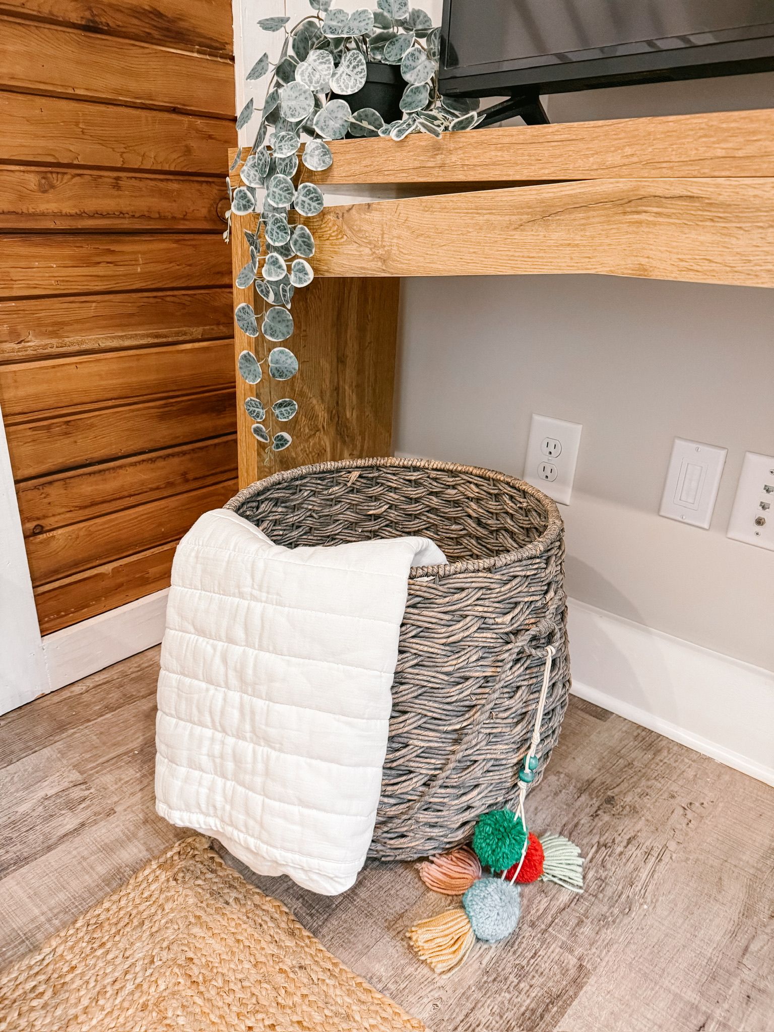 A corner of a room with a woven basket, a white quilted blanket, and a small rug on the floor.