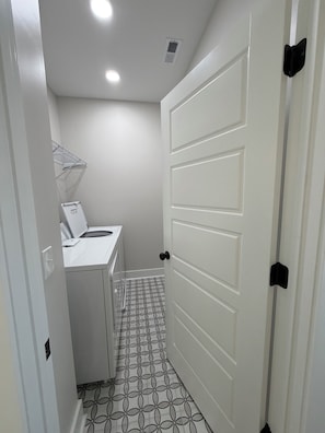 A narrow hallway with a white door, a washing machine, and a shelf with a white appliance. The floor has a geometric tile pattern. - A modern relaxed retreat in charming historic downtown Springfield! (Springfield)