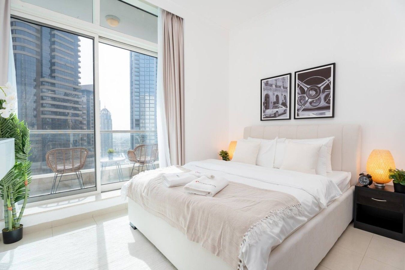 Una habitación de hotel moderna con una cama grande, dos lámparas de noche, un ventanal con vistas a la ciudad y dos cuadros enmarcados en la pared.