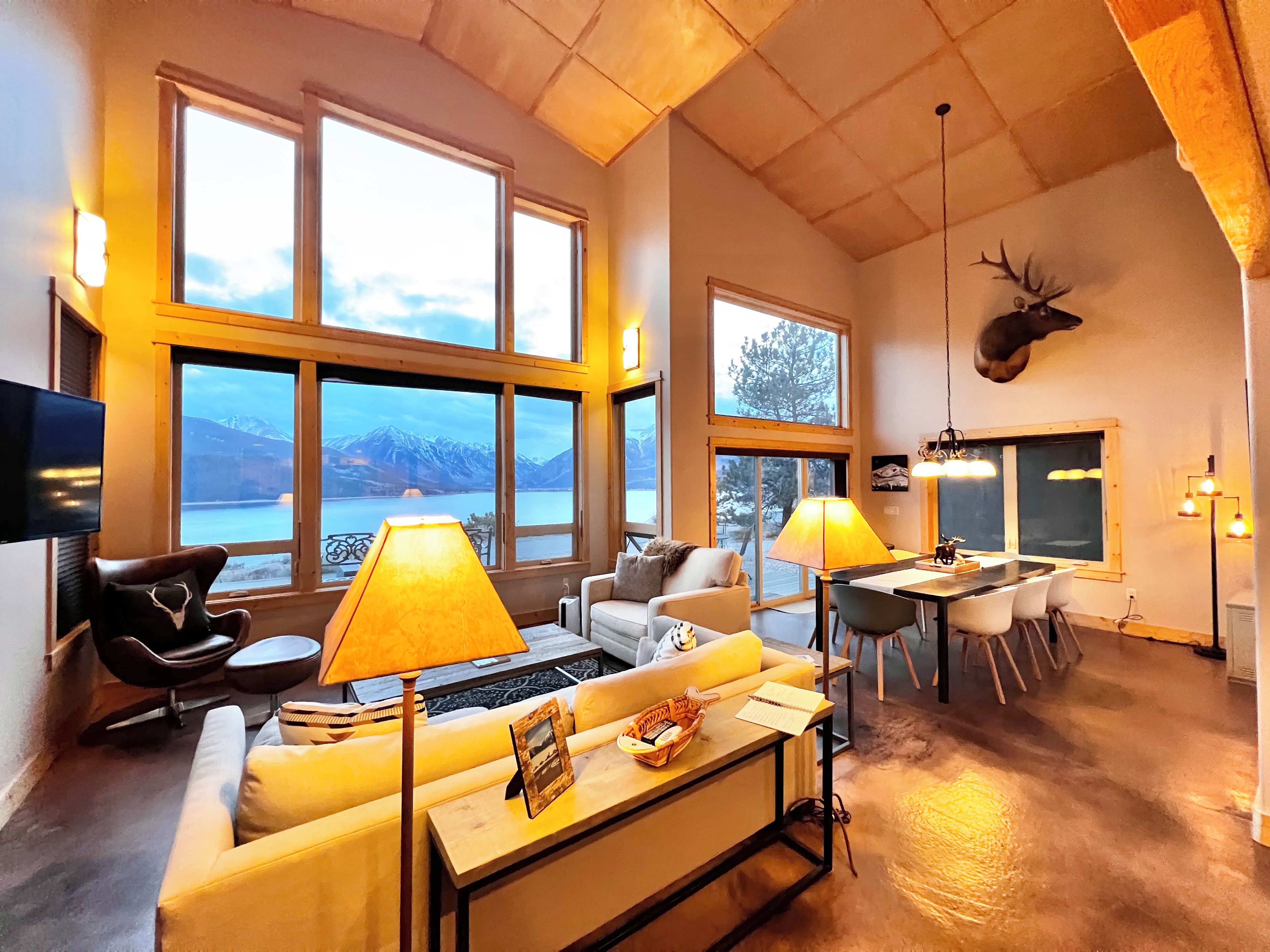 A spacious living area with a large window overlooking a mountainous landscape, a dining area with a table and chairs, and a mounted animal head on the wall.