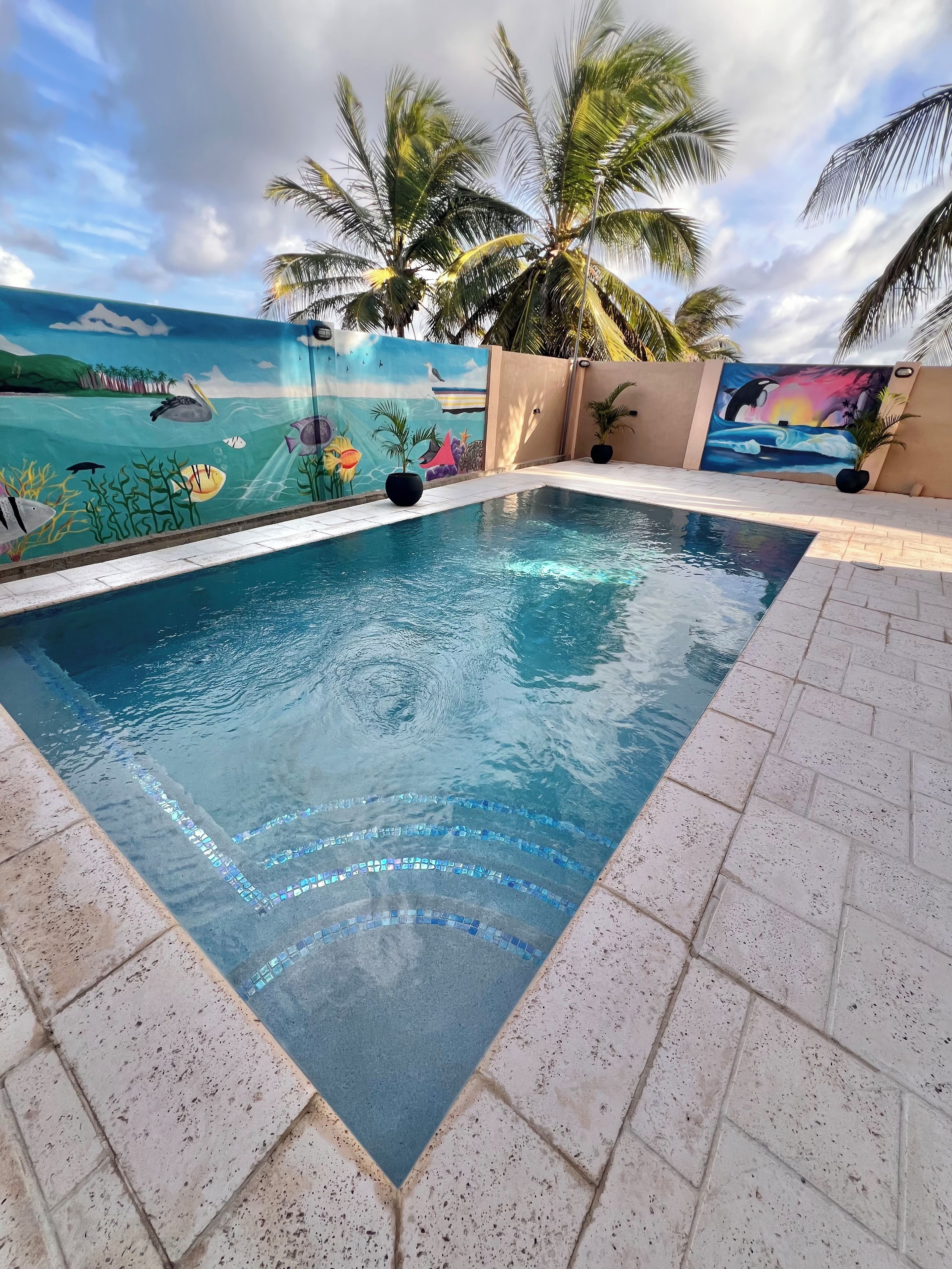 Une piscine dont les bords sont recouverts de carreaux, entourée de palmiers et ornée d’une fresque représentant des scènes sous-marines.